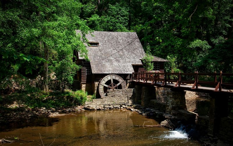 A rustic wooden watermill with a shingled roof sits by a stream, surrounded by dense green trees. Featured in many Old Mill Pigeon Forge guides, this scenic spot has a wooden bridge and sunlight filtering through the foliage.
