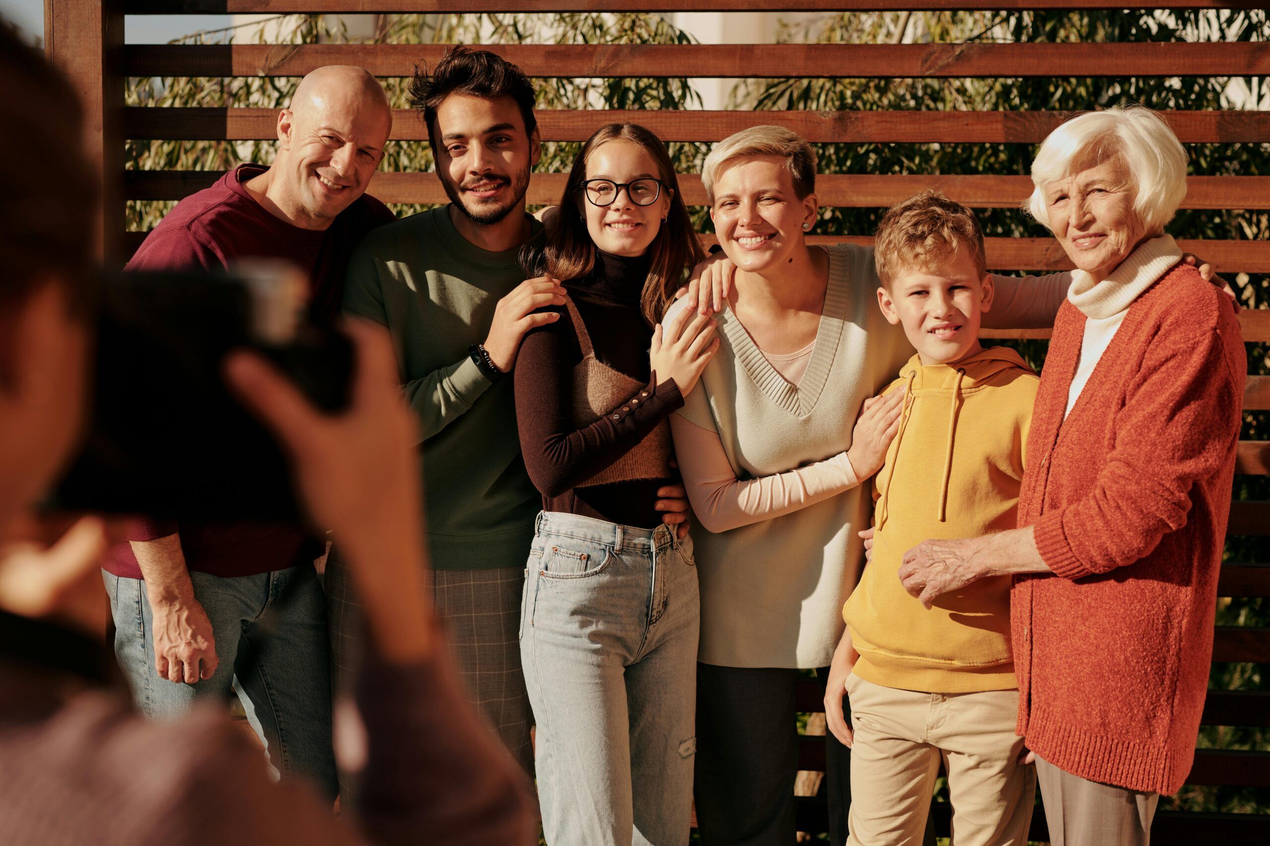A group of six people of different ages stand close together, smiling for a family reunion photo outdoors. One person in the foreground is taking their picture. Trees and wooden slats are visible in the background, evoking the charm of the Smoky Mountains.