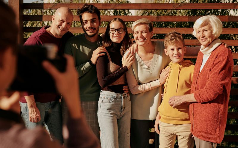 A group of six people of different ages stand close together, smiling for a family reunion photo outdoors. One person in the foreground is taking their picture. Trees and wooden slats are visible in the background, evoking the charm of the Smoky Mountains.