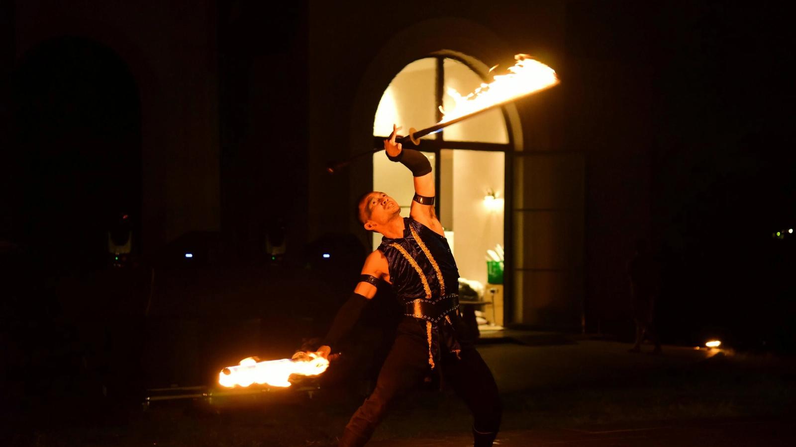 A fire performer in dark clothing dramatically poses at night, holding two flaming batons—one above his head and one extended outward—against the backdrop of an arched doorway, reminiscent of the thrilling Dinner Shows in Pigeon Forge.