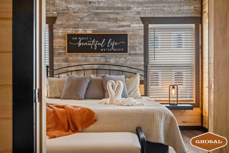 A cozy bedroom in lodging near Gatlinburg features a metal bed frame, neutral bedding, an orange blanket, and two towel swans. Sunlight streams through window blinds beneath a sign reading, “Oh what a beautiful life we’ve built.”.