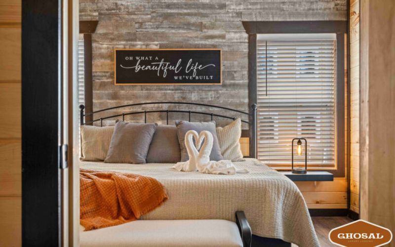 A cozy bedroom in lodging near Gatlinburg features a metal bed frame, neutral bedding, an orange blanket, and two towel swans. Sunlight streams through window blinds beneath a sign reading, “Oh what a beautiful life we’ve built.”.