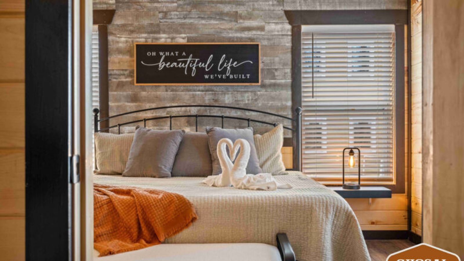 A cozy bedroom in lodging near Gatlinburg features a metal bed frame, neutral bedding, an orange blanket, and two towel swans. Sunlight streams through window blinds beneath a sign reading, “Oh what a beautiful life we’ve built.”.