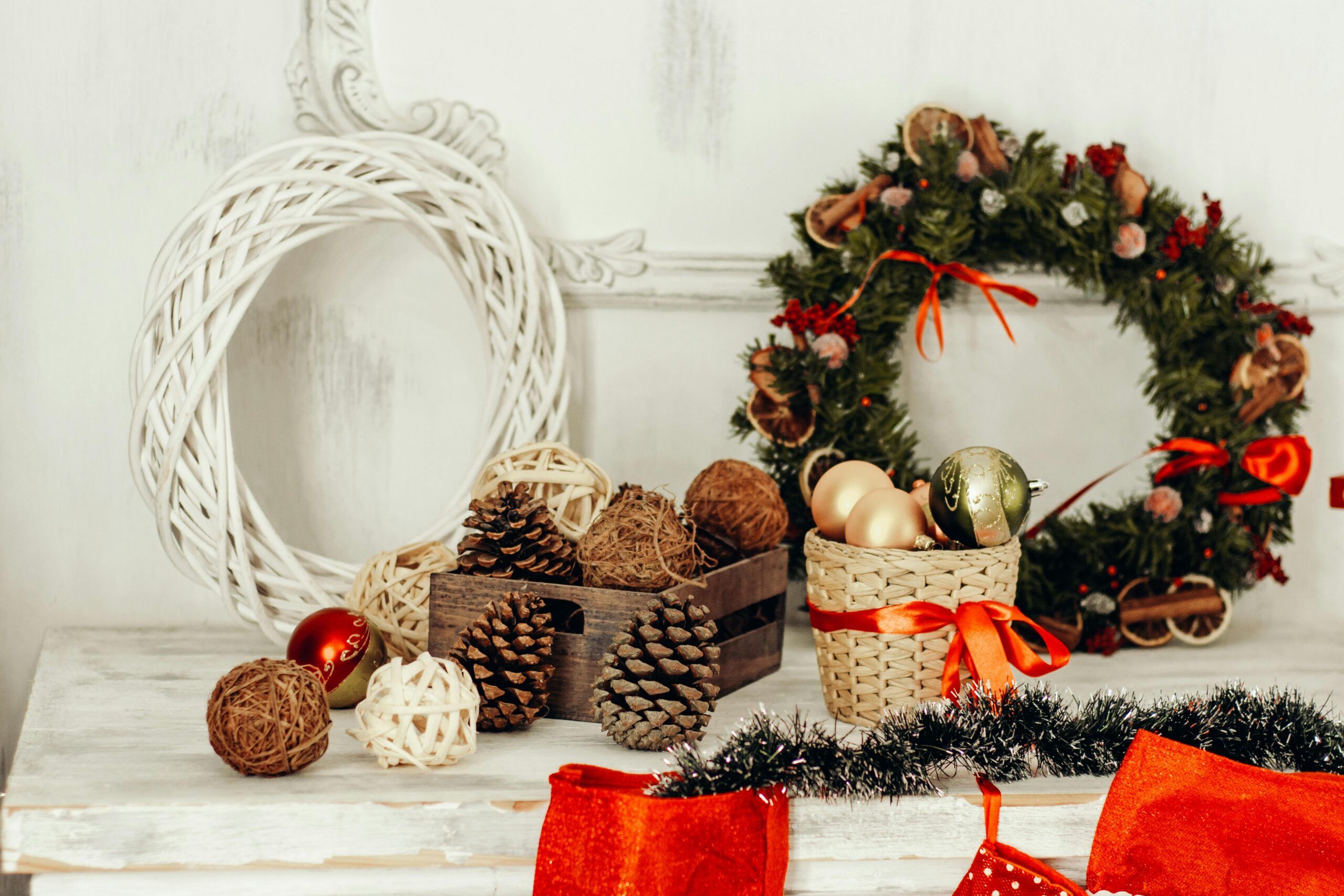 A white table decorated with pinecones, wicker balls, and Christmas ornaments in baskets evokes the charm of Gatlinburg Christmas, set next to a green holiday wreath with red ribbons and white wicker wreath, with red stockings hanging below.