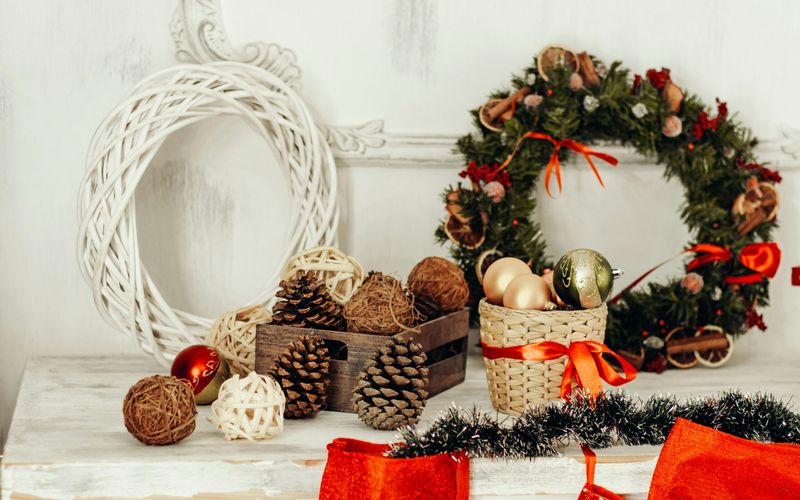 A white table decorated with pinecones, wicker balls, and Christmas ornaments in baskets evokes the charm of Gatlinburg Christmas, set next to a green holiday wreath with red ribbons and white wicker wreath, with red stockings hanging below.