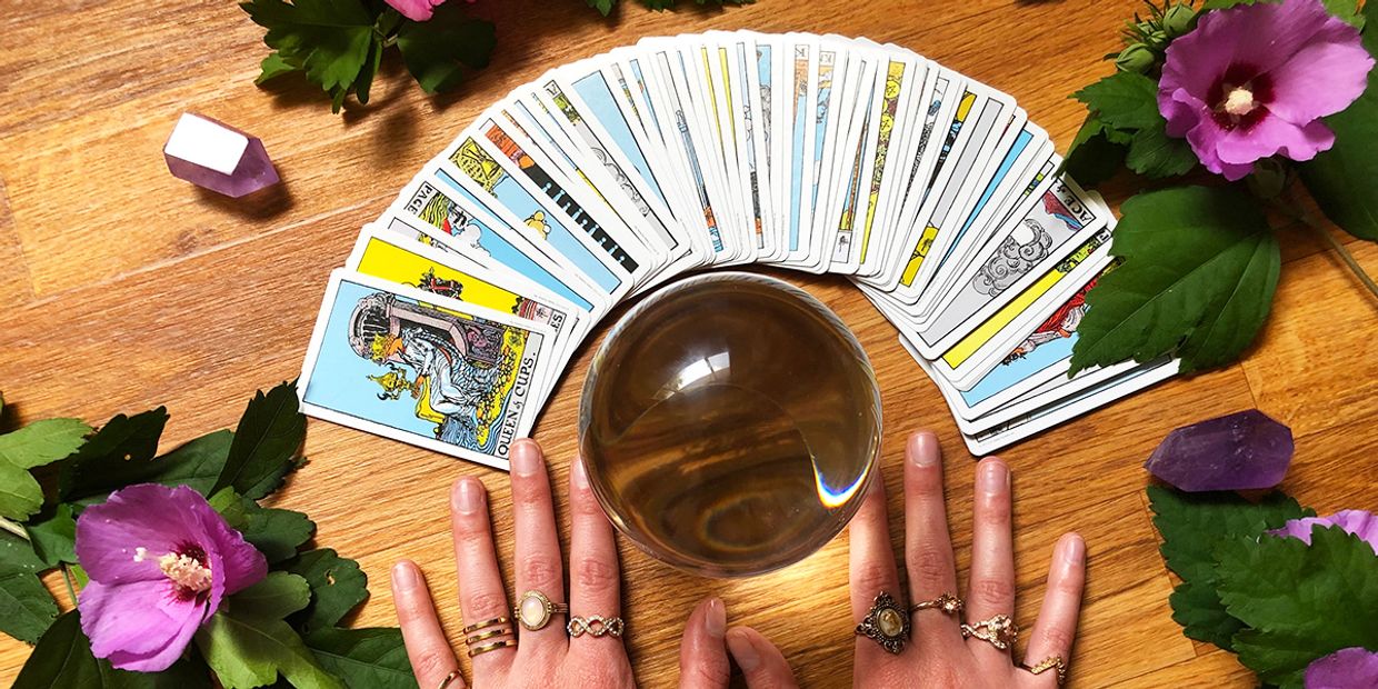an image showing a woman with both hands flat on a wooden desk, surrounded by a deck of tarot cards, spread out and a crystal ball sitting in the centre.