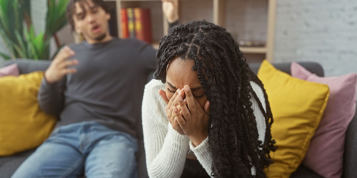 A couple arguing on a grey sofa, both looking upset.