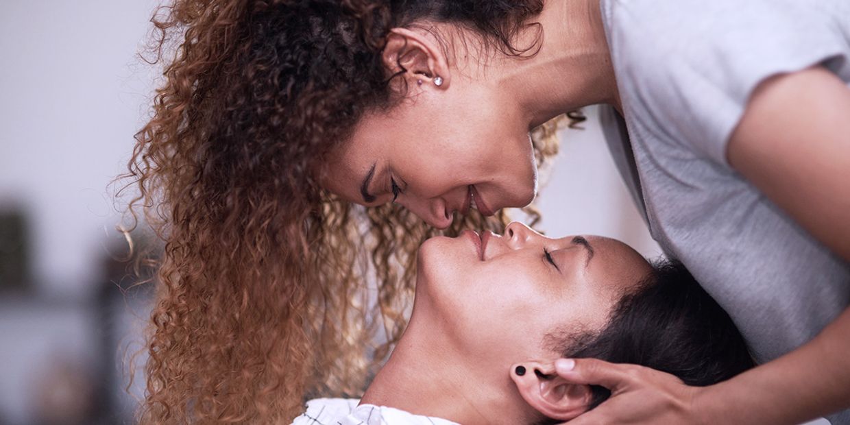 2 Lesbians in love, one is lying down and the other is leaning over her planting a kiss on her lips.