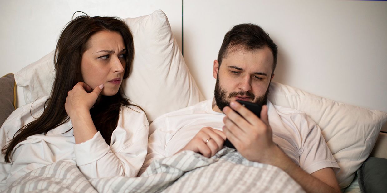 Couple in bed; woman looks concerned while man focuses on phone.