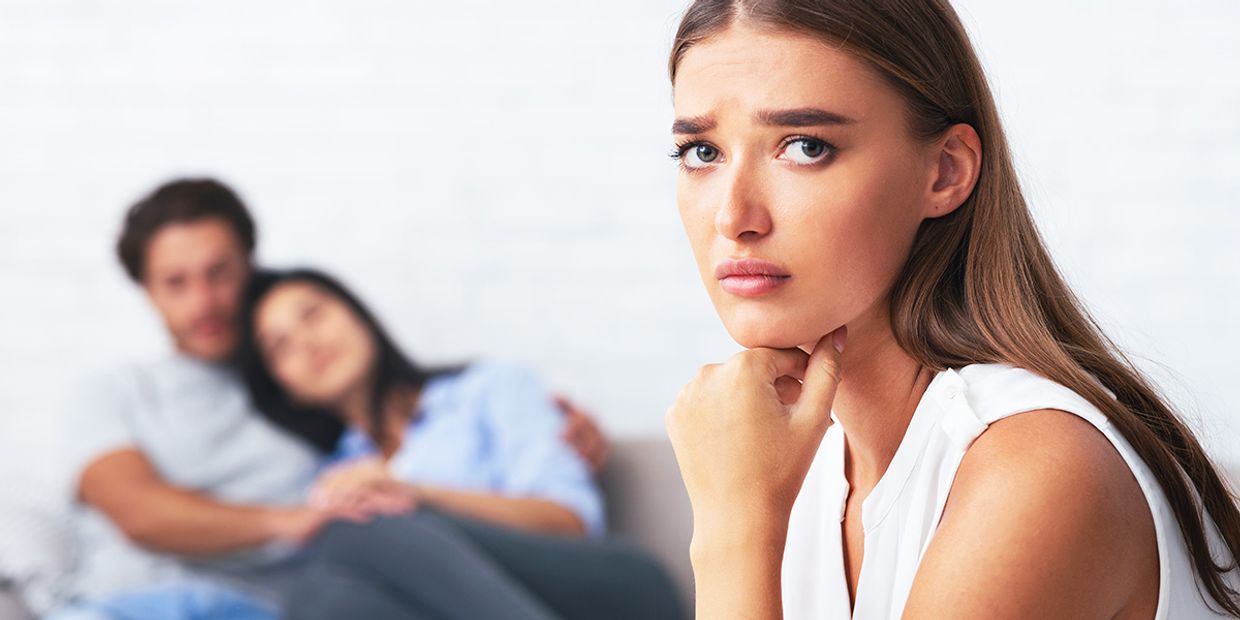 A sad woman looking into the camera with a couple sitting together in the background.