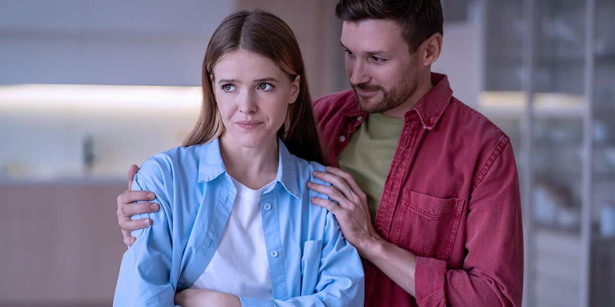 A male and female couple in their home. The woman looks apprehensive, whilst the man tries to embrace her.