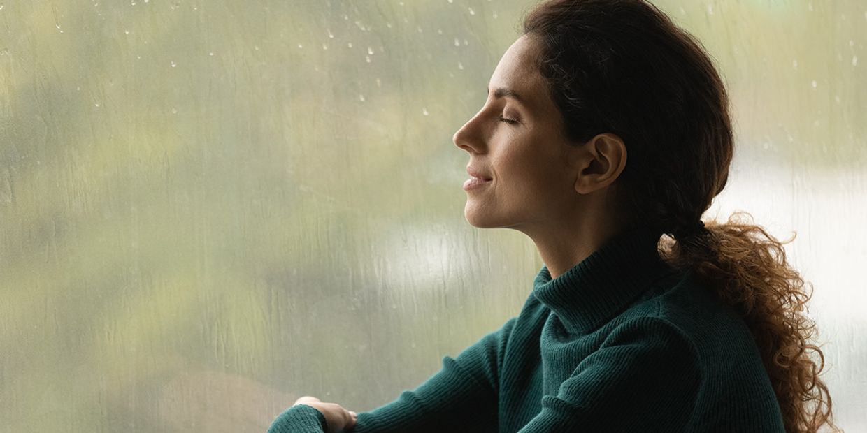 A middle aged woman sitting by a window, it's raining outside, she is peaceful.