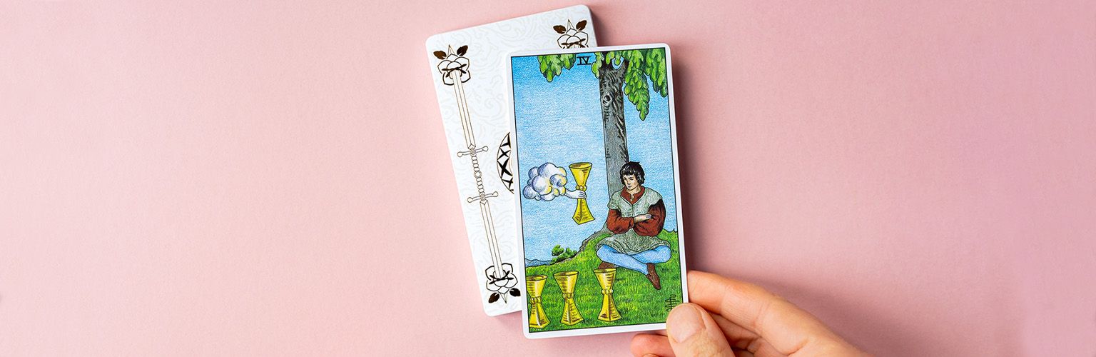 A Four of Cups tarot card being held over a soft pink background.