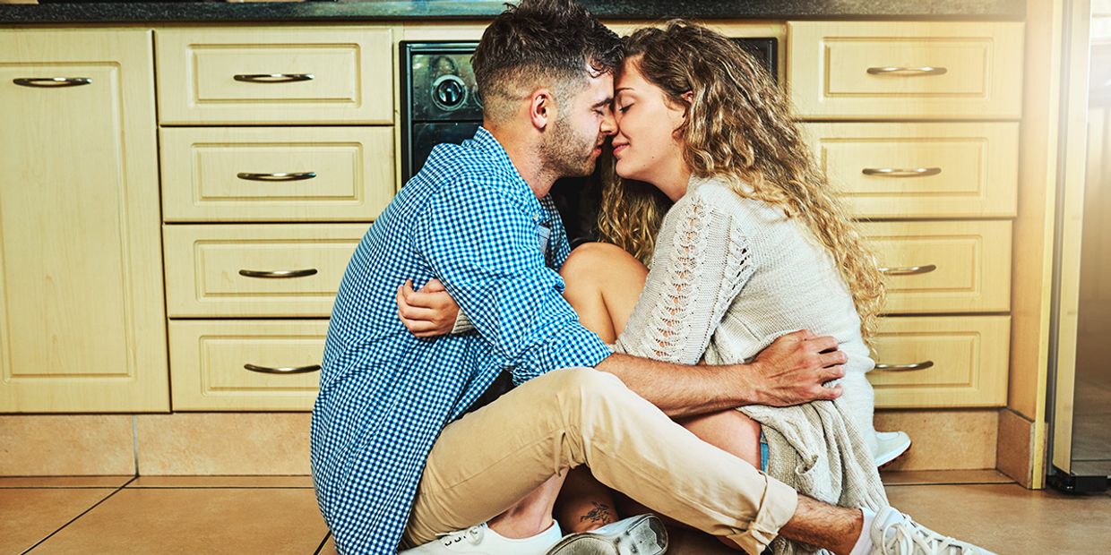 A cute young couple sitting on the floor of a kitchen, holding each other and looking into each other's eyes for an intimate moment together.