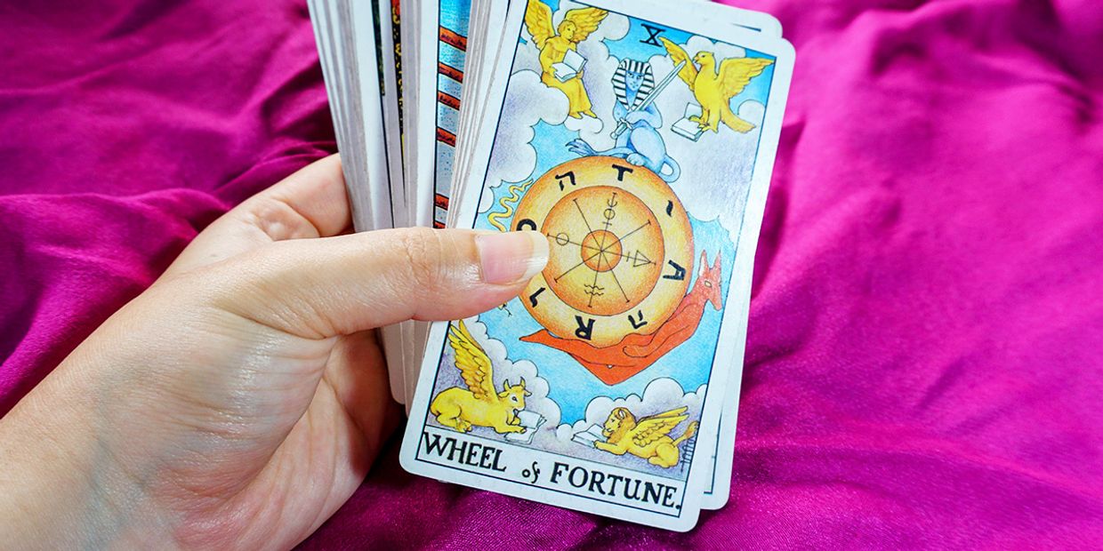 A female hand holding a deck of tarot cards with the Wheel of Fortune card facing up against a vibrant magenta cloth, symbolising destiny, life cycles, and spiritual guidance.