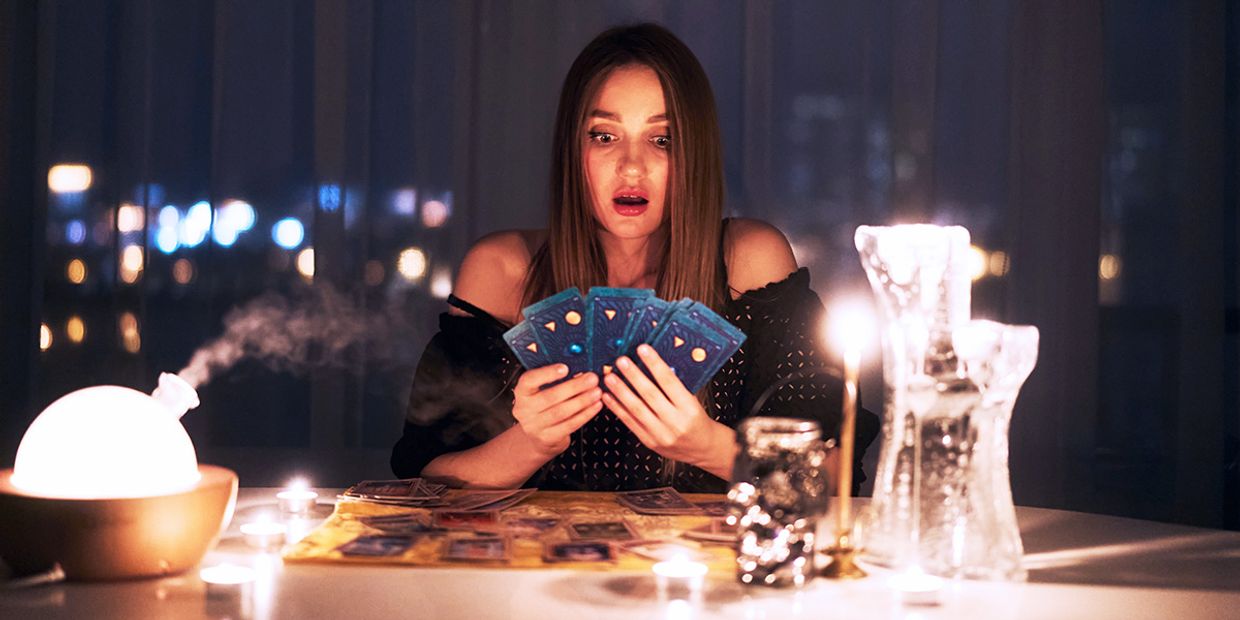 A surprised looking female tarot card reader, holding a spread of tarot cards in a darkly lit living room.