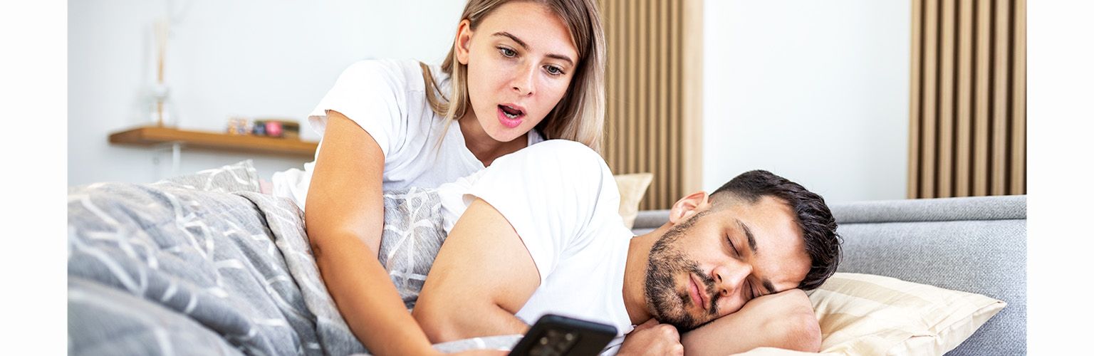 Woman shocked by man's phone in bed, revealing potential cheating signs.