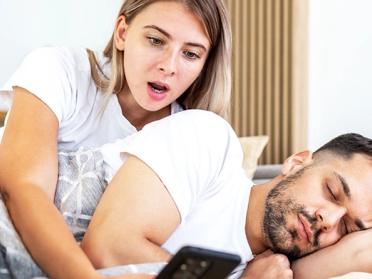 Woman shocked by man's phone in bed, revealing potential cheating signs.