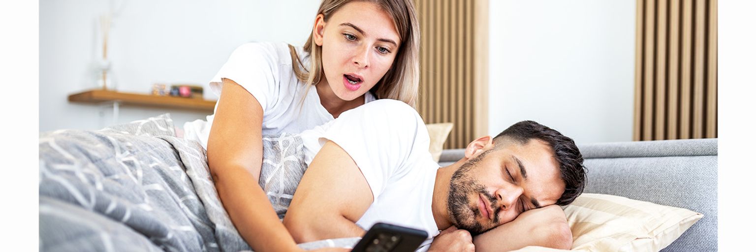 Woman shocked by man's phone in bed, revealing potential cheating signs.