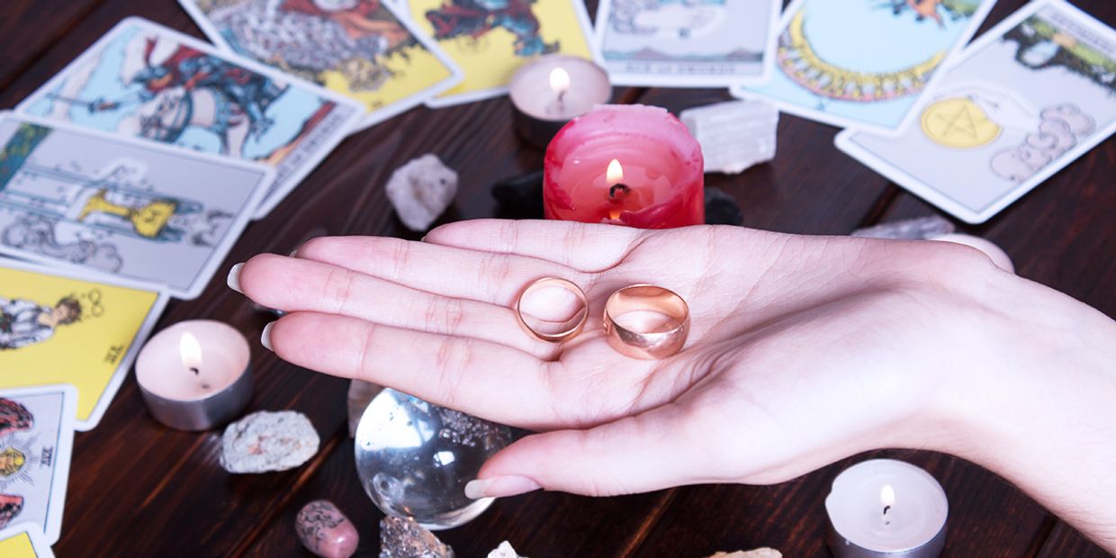 Hand holding two wedding rings surrounded by tarot cards, candles and crystals, making a love tarot reading.