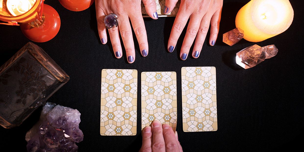 Hands over three tarot cards face down with candles, crystals and other spiritual items around, as part of a three-card tarot spread.