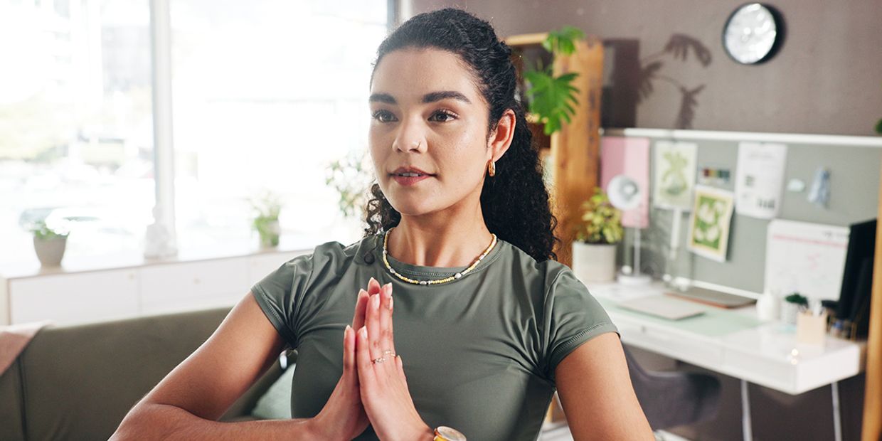 A young woman standing in a meditating pose, in her office.