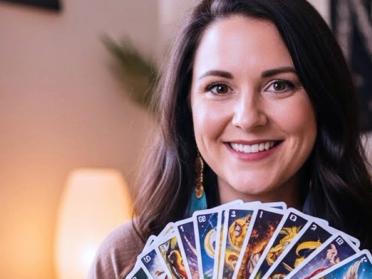 Smiling woman holding a fan of colourful tarot cards in a warm and spiritual room with candles and decor, symbolising intuitive psychic readings and positive customer experiences.