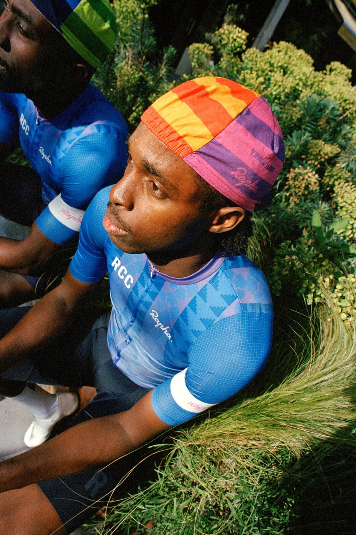 Cyclist in blue jersey and multicolored cap sitting on grass during an outdoor race moment