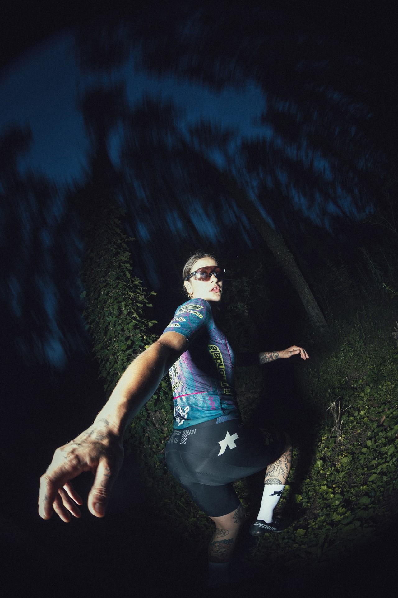 Cyclist in a blue jersey and sunglasses rides hard at night, arms out in a dramatic motion