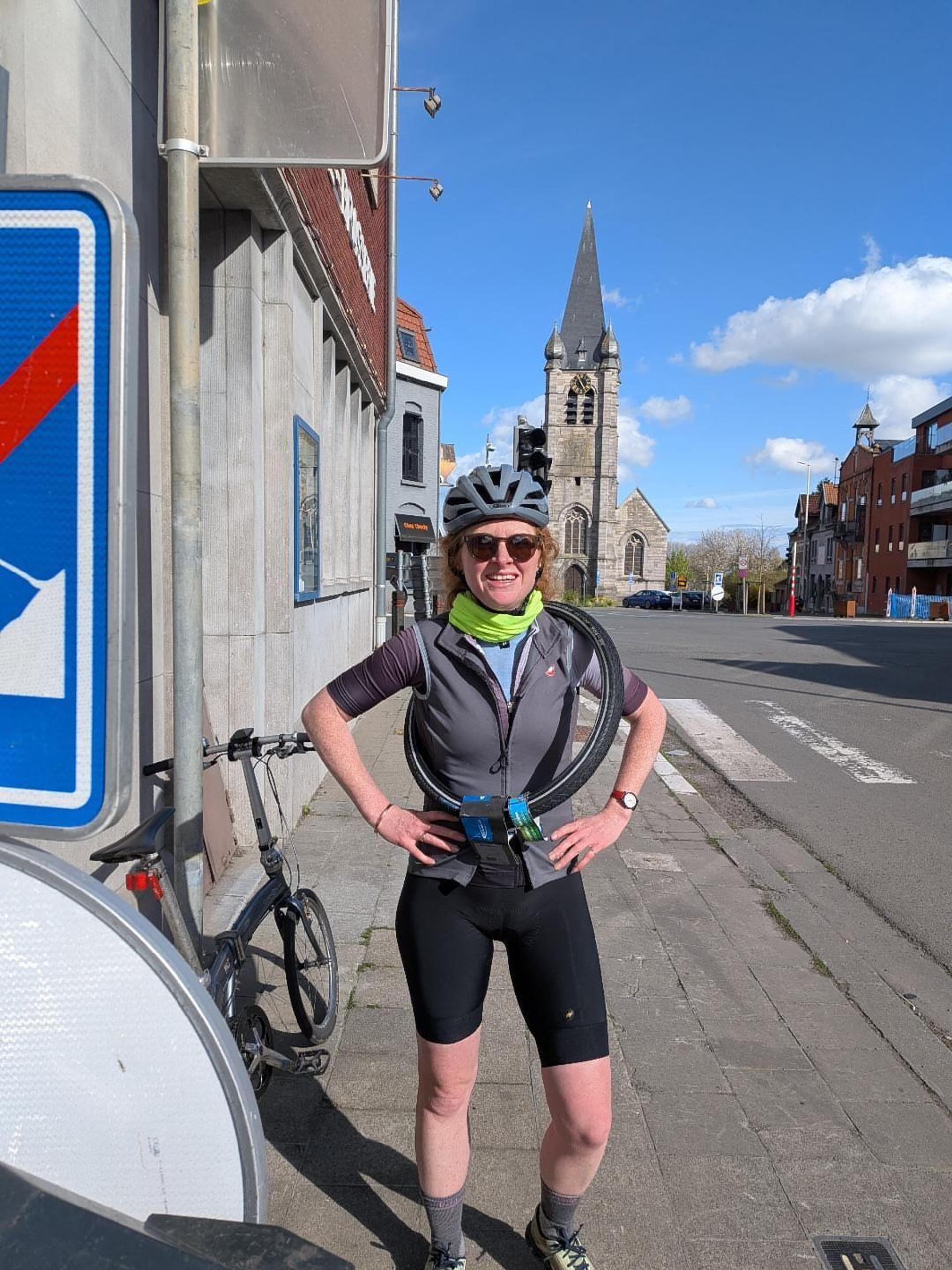 Armorel Jackson poses with a spare tyre slung around her shoulders and a folding Brompton bike propped against a sign post in a Belgian town, with a church spire in the background