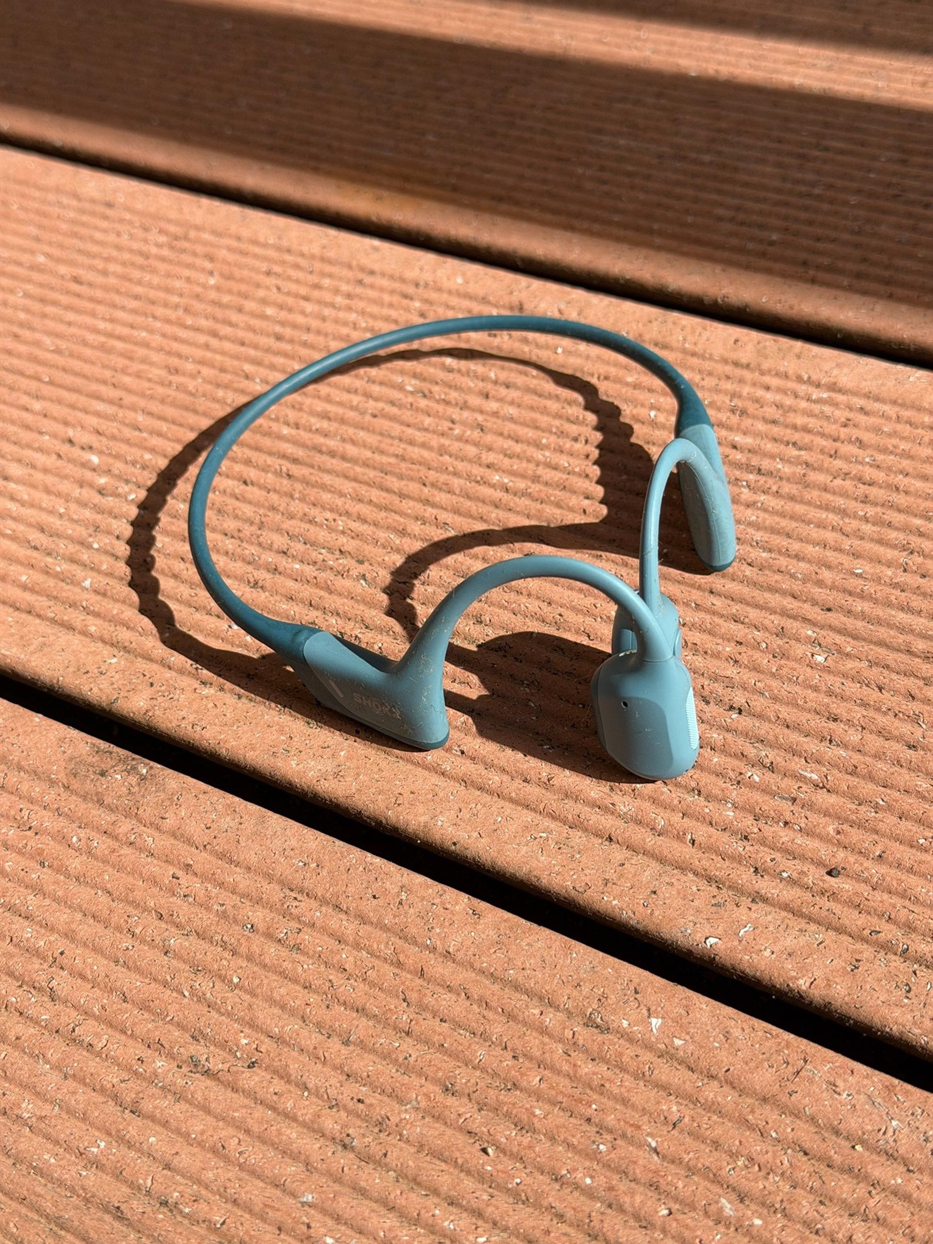Teal Shokz bone-conduction headphones resting on a sunlit wooden deck.