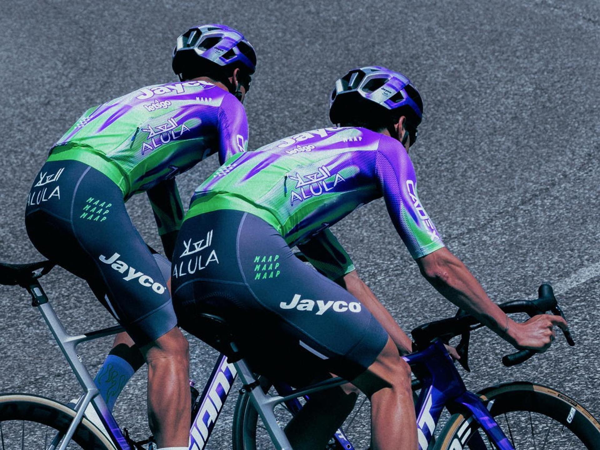 Two cyclists in green-purple kits ride closely together on a road, hands at the handlebars