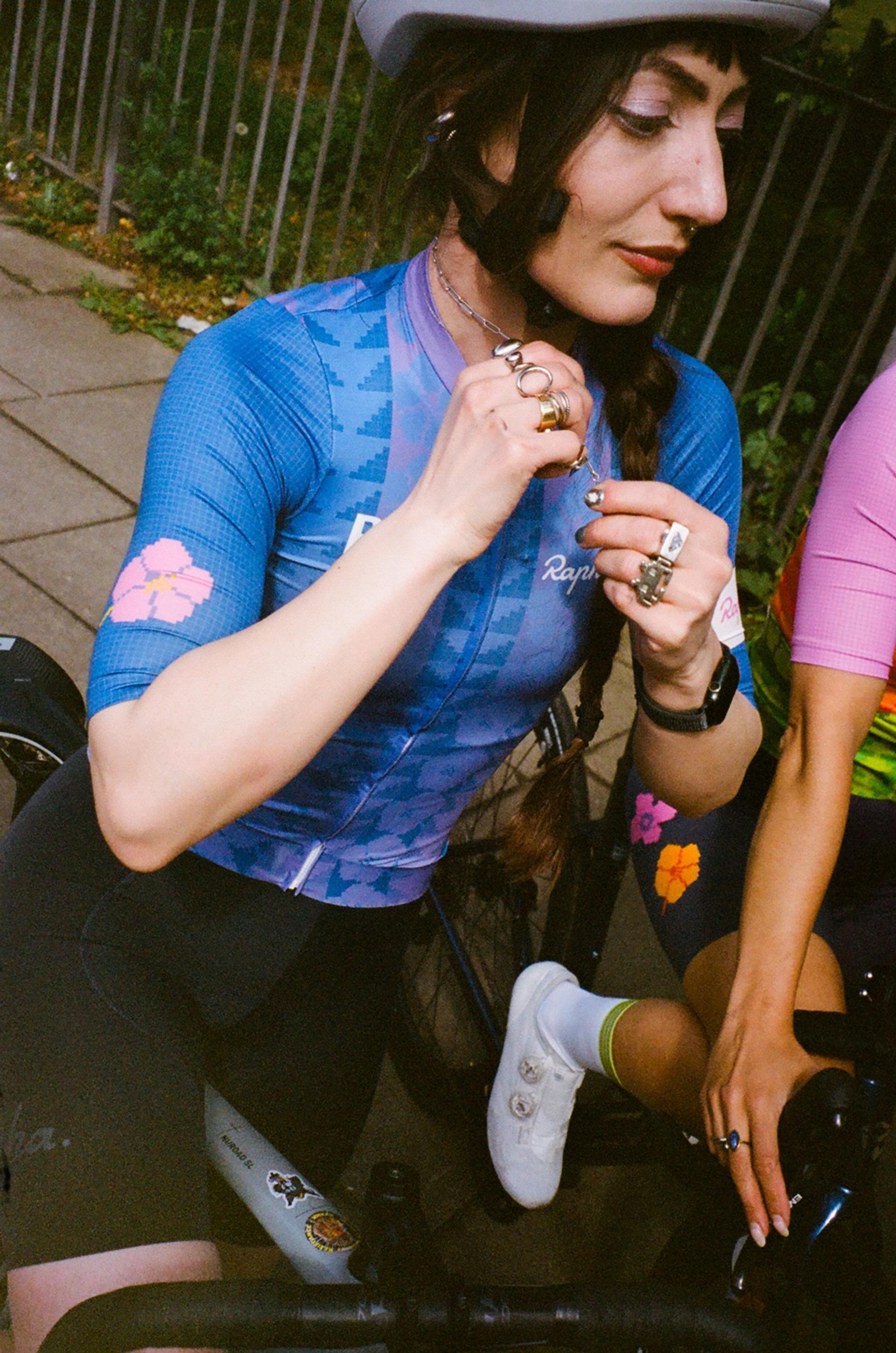 Cyclist in blue jersey adjusts a watch while sitting outdoors near a railing, holding a drink
