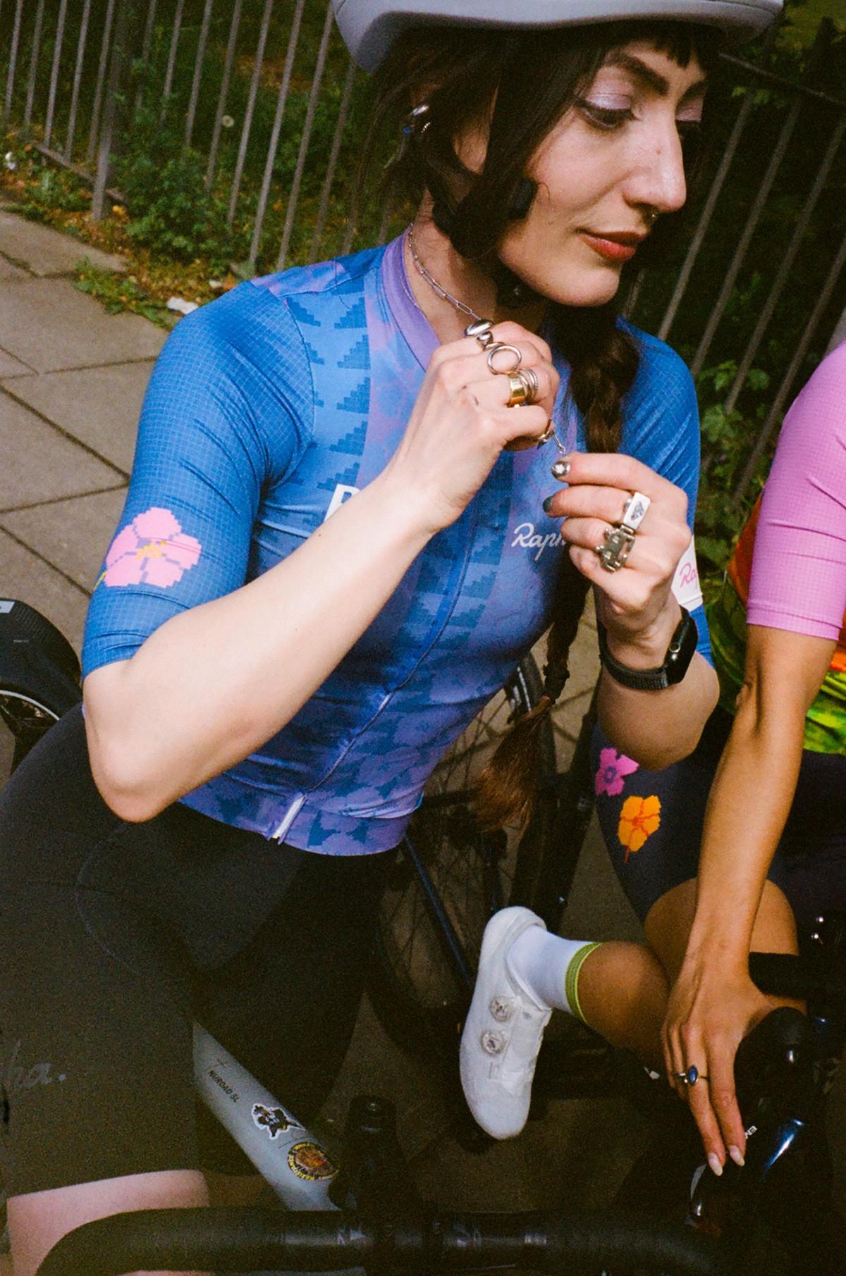 Cyclist in blue jersey adjusts a watch while sitting outdoors near a railing, holding a drink