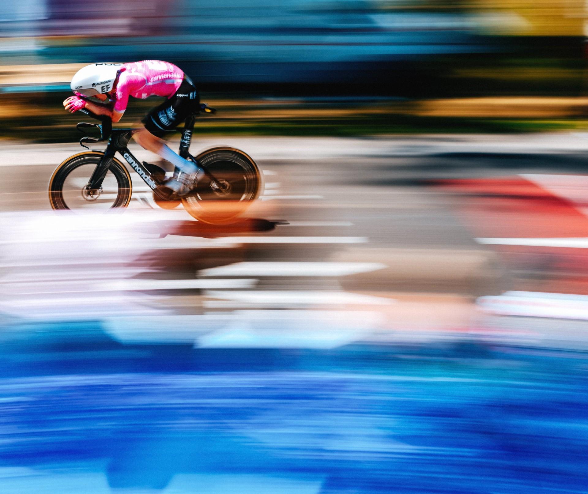 Time-trial cyclist in a pink jersey and aerodynamic helmet rides a bike, captured with strong motion blur.