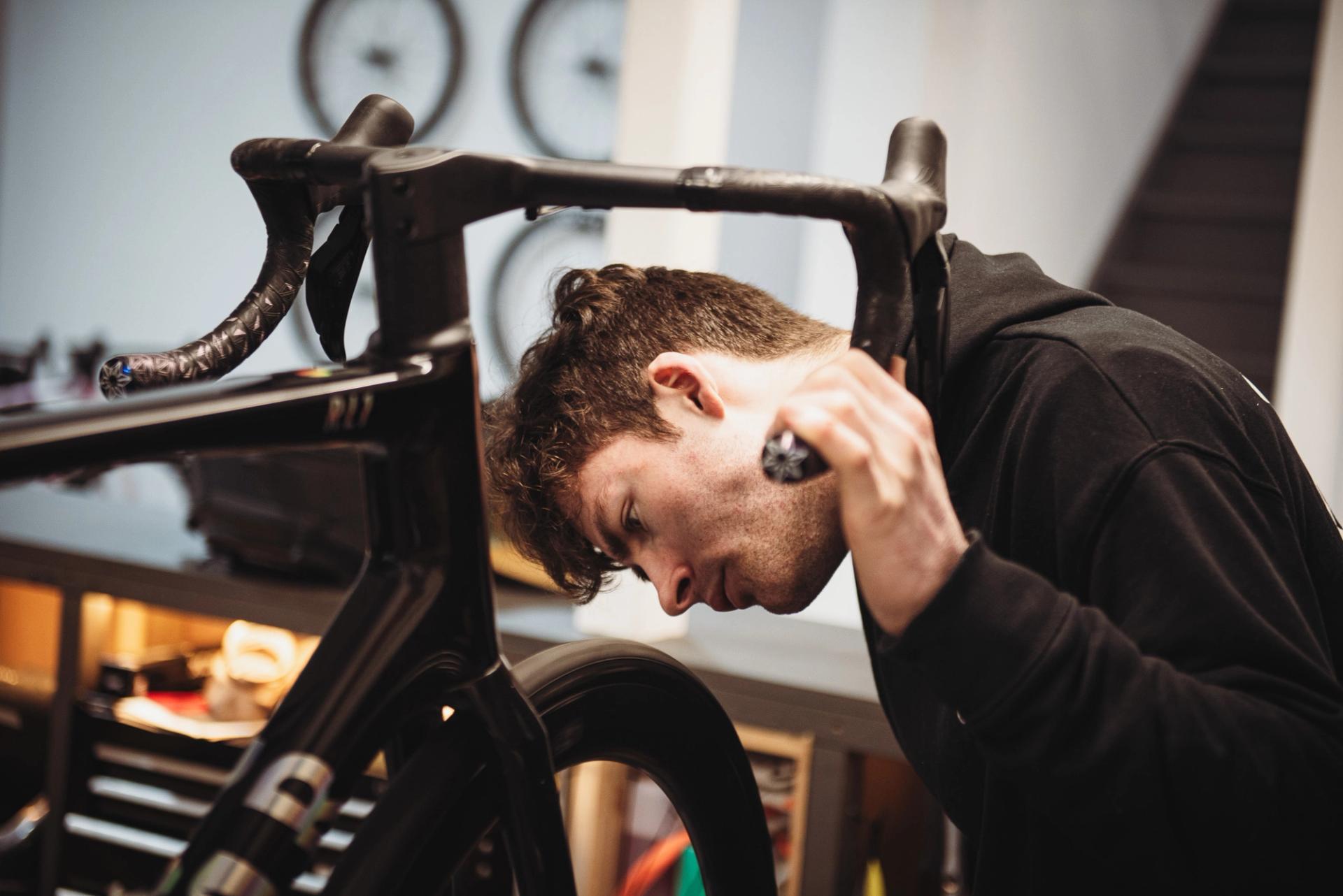 Close-up of a bicycle mechanic inspecting the front wheel and cockpit of a road bike, leaning in closely while holding a tool in a workshop setting.
