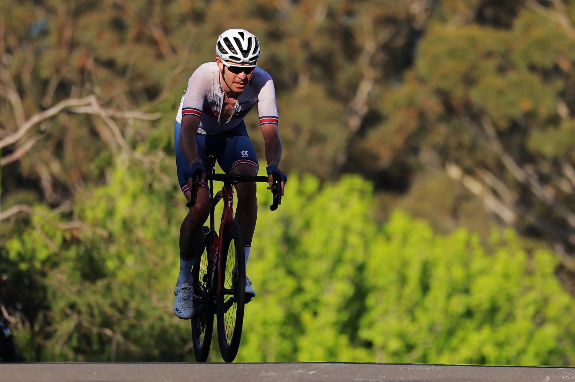 Cyclist in a helmet and sunglasses rides a road bike along a tree-lined road.