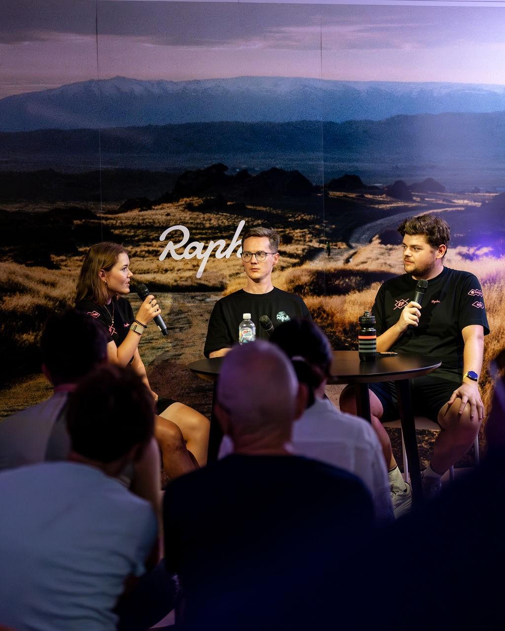 Three people sit on stage holding microphones during a Rapha event in a scenic landscape backdrop.
