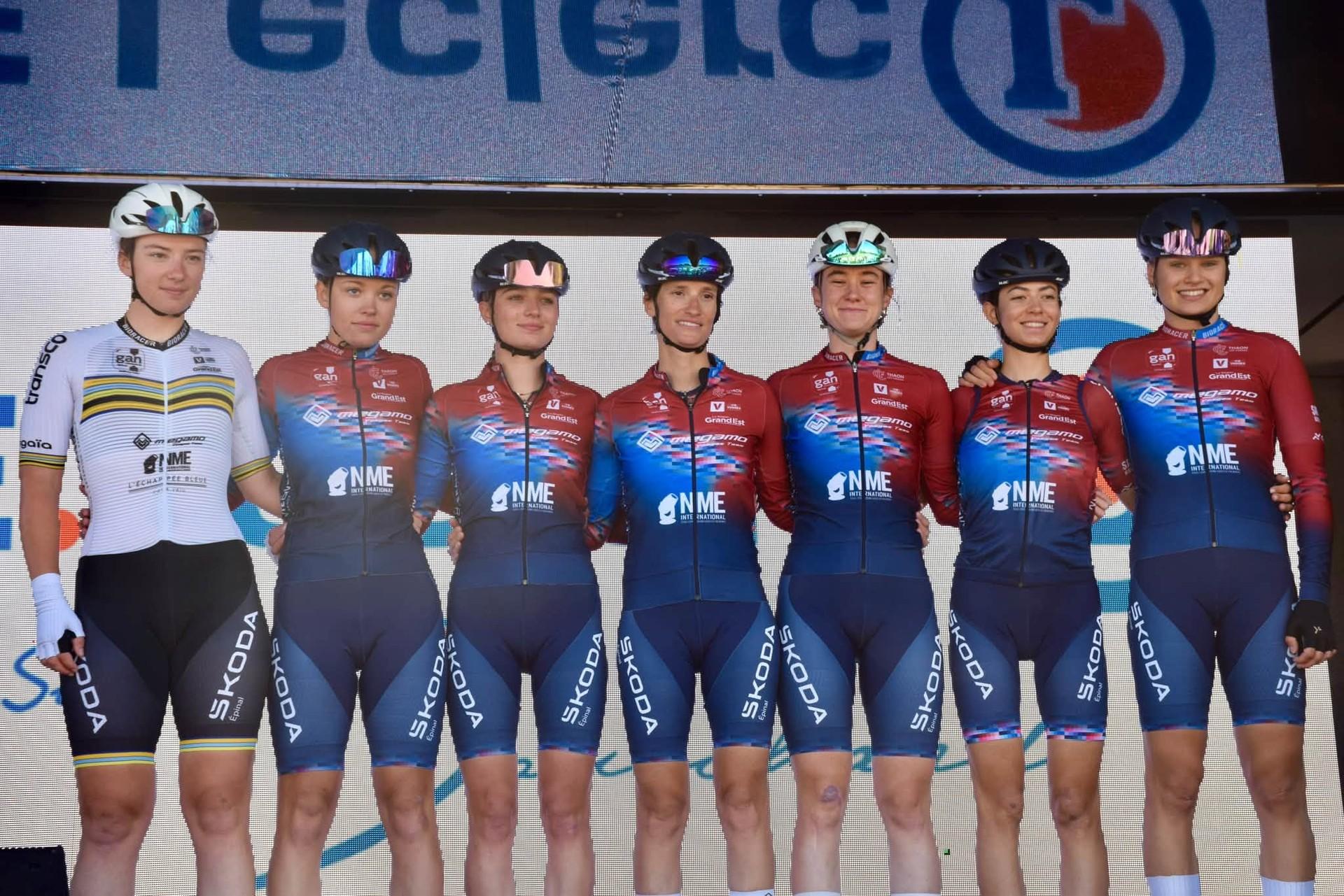 Grace Reynolds and six Megamo Vosges teammates pose in cycling kits beneath a sponsor sign.