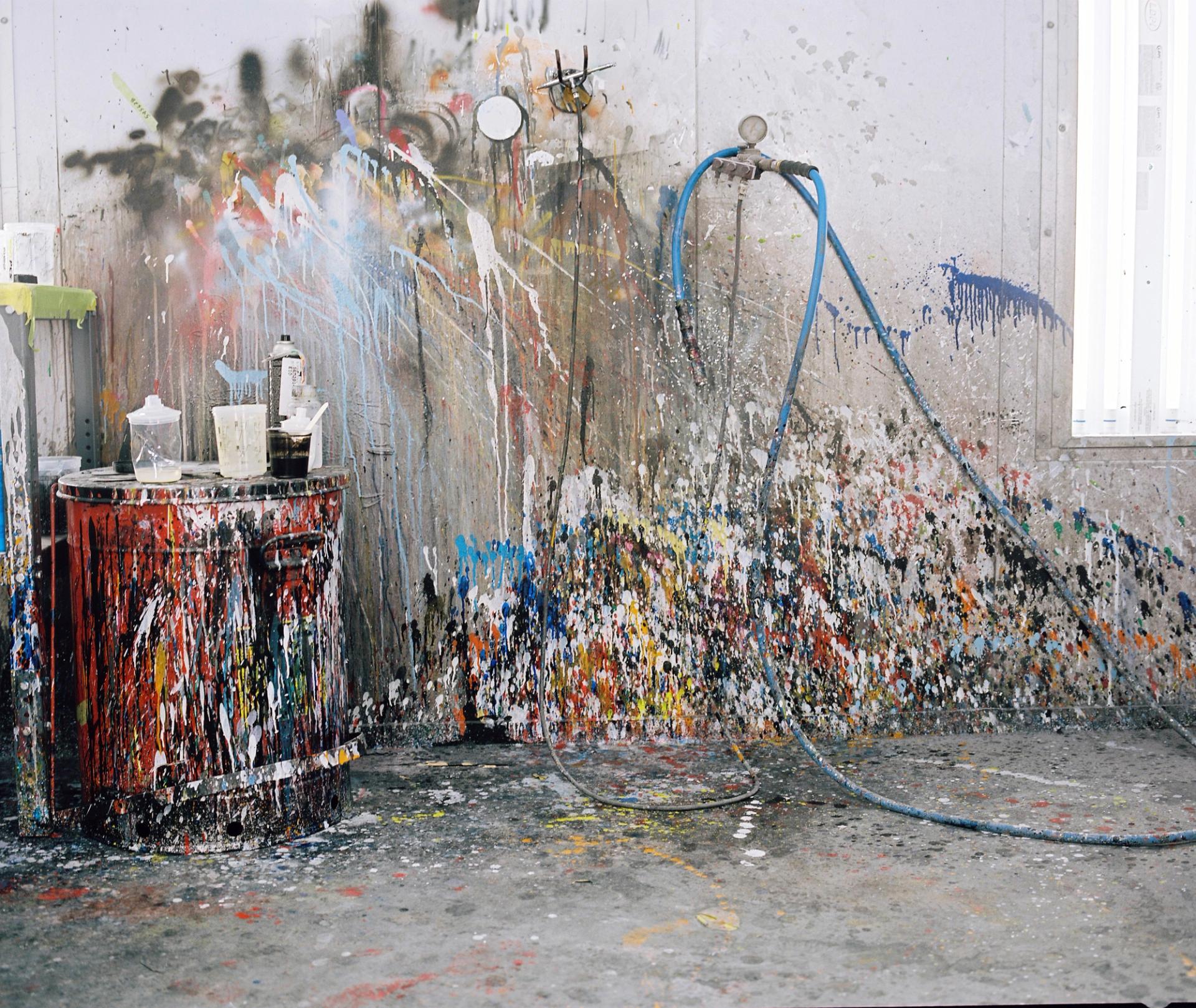 A wider view of the paint booth workspace, layers of multicoloured overspray creating an abstract patina on every surface—walls, hoses, and fixtures alike.