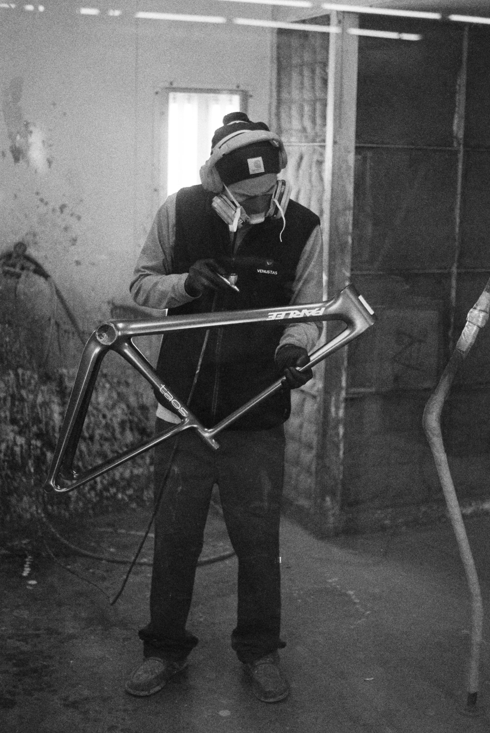 Black and white portrait of a Parlee team member standing with a completed bicycle frame, holding it at arm's length as if presenting their work.