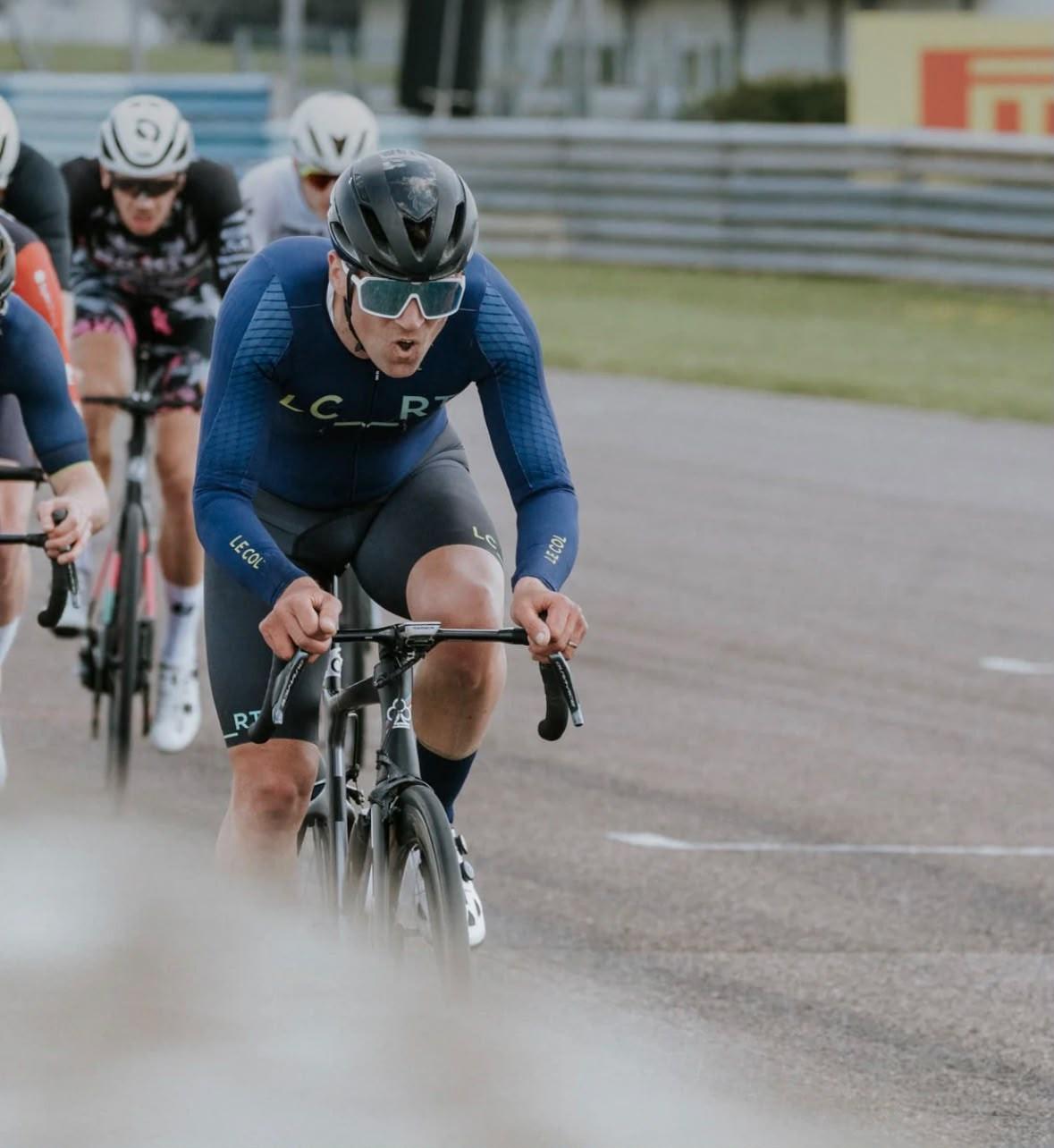 Cyclist in blue kit leads a road race pack on a paved circuit, drafting during a sprinted chase.