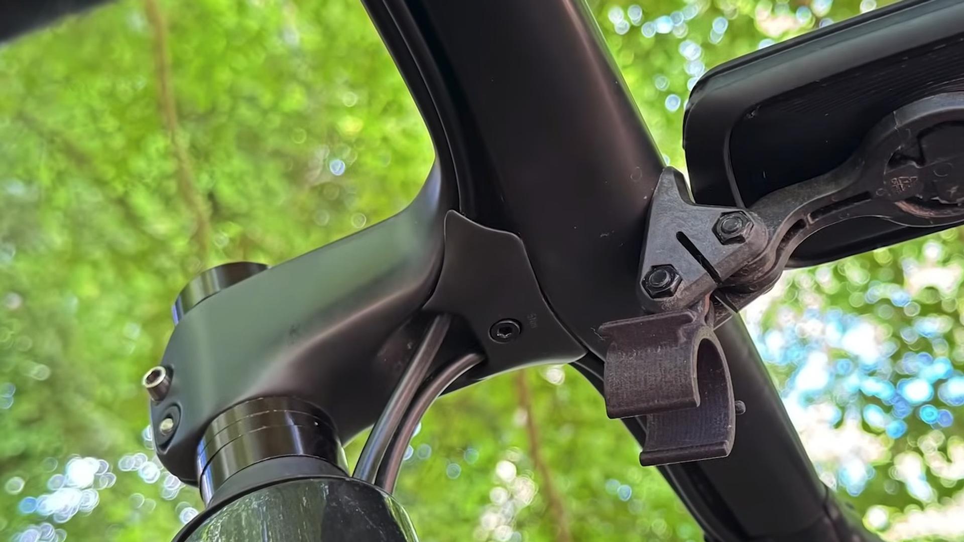 Integrated carbon bicycle handlebars with 3D-printed accessory mount and computer out-front display against green foliage.