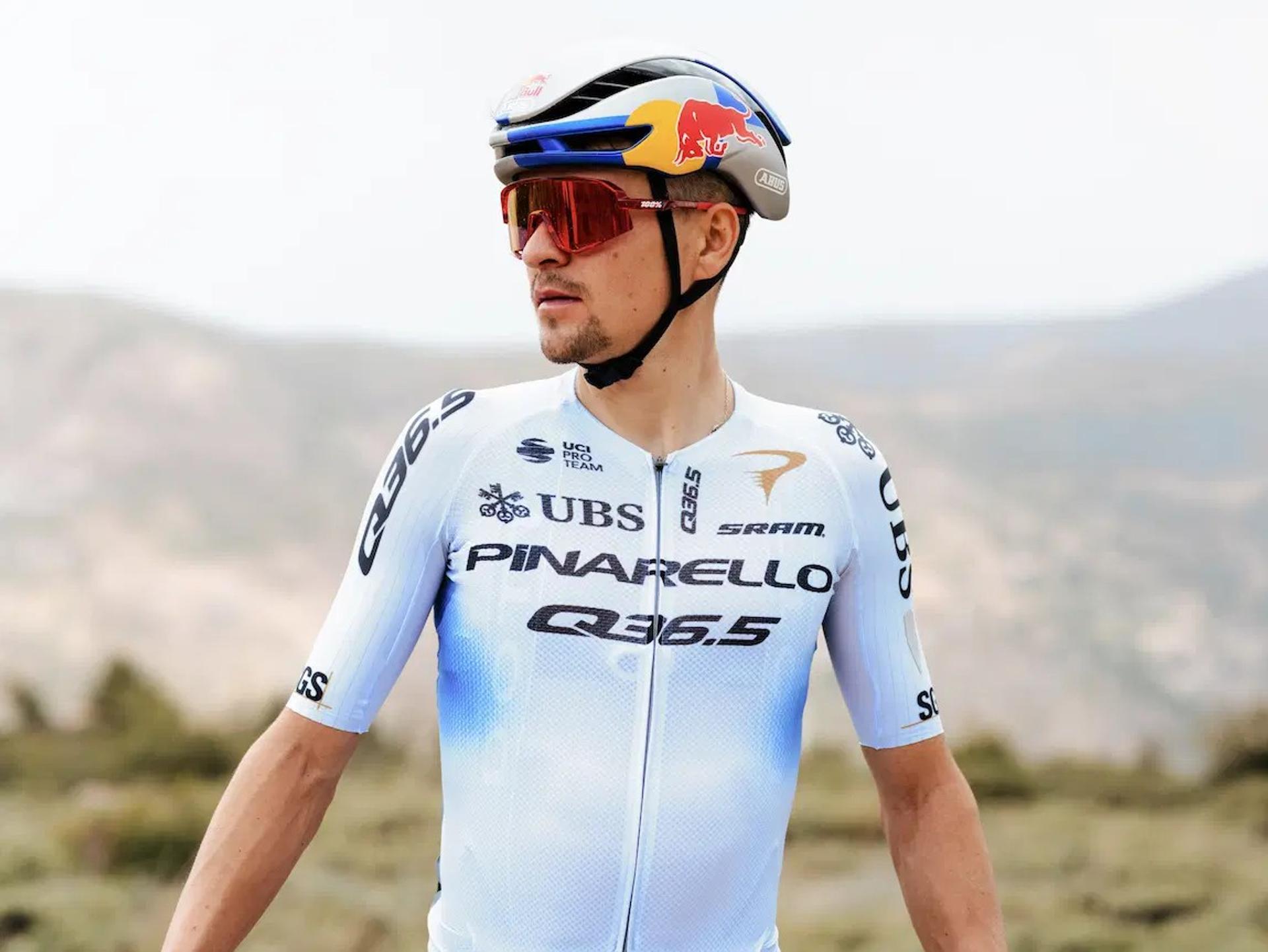 Cyclist in PBAREYLO team kit stands outdoors, wearing sunglasses and helmet with race numbers