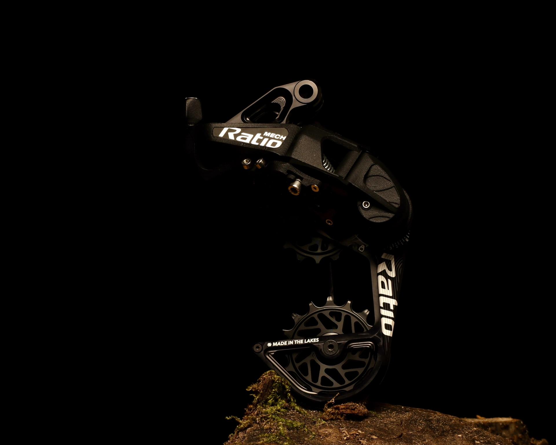 A black Ratio Mech rear derailleur photographed against a completely black background, standing upright on a small patch of mossy ground with the words “Made in the Lakes” visible on the lower cage.