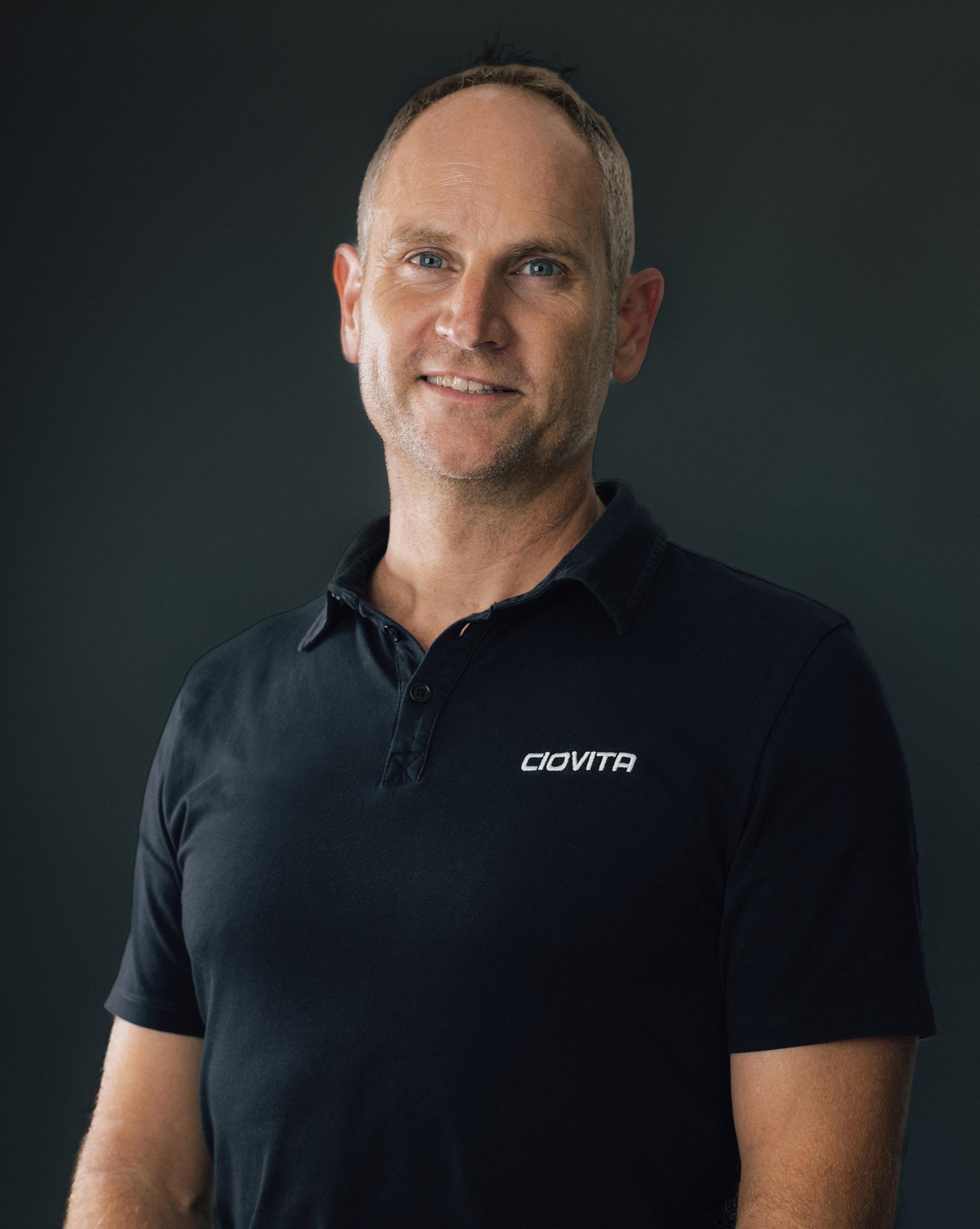 Portrait of a smiling man in a dark “CIOVITA” polo shirt against a dark background.