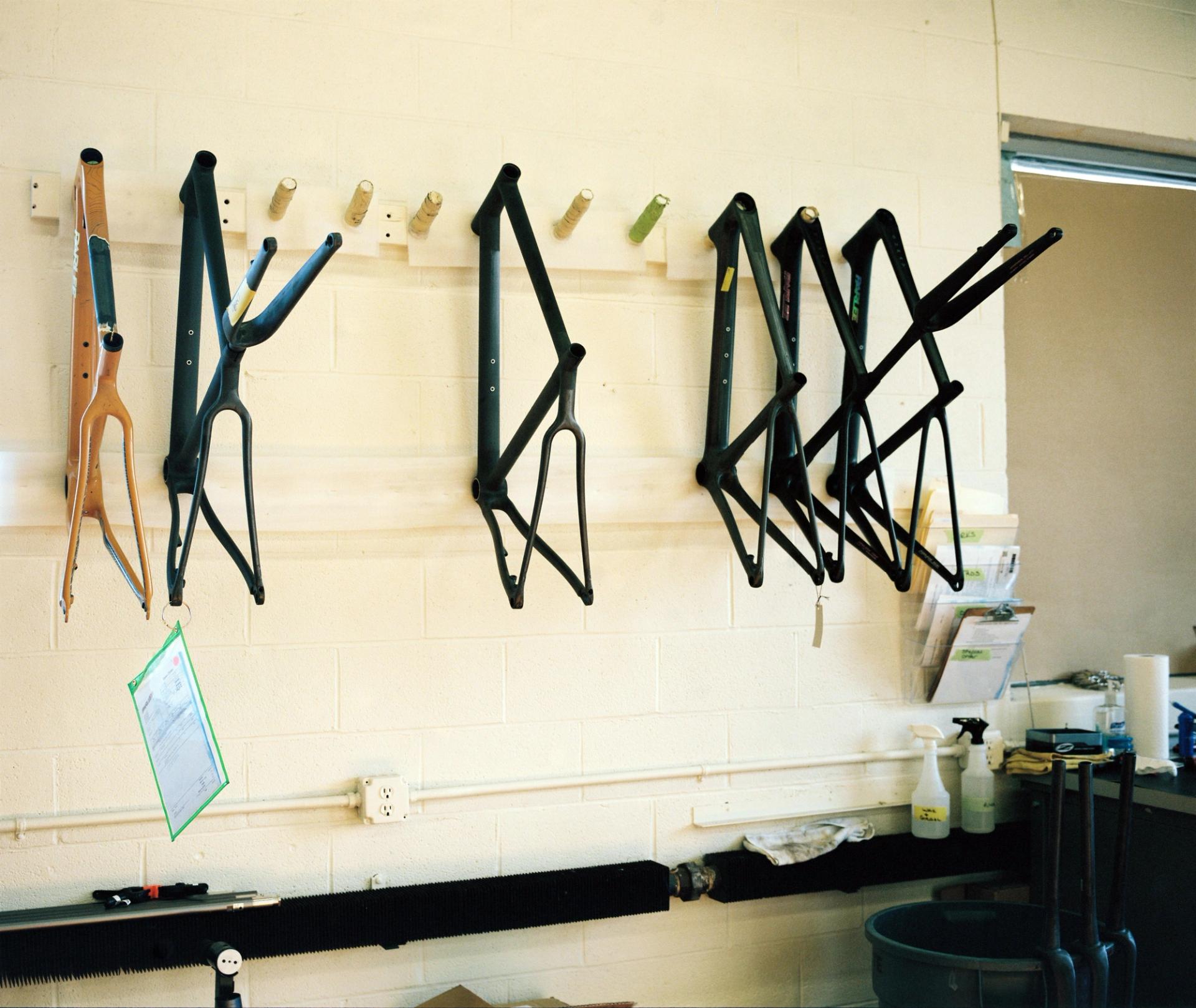 Carbon fibre bicycle forks hung on a workshop wall, their black forms creating angular silhouettes against a pale yellow background.
