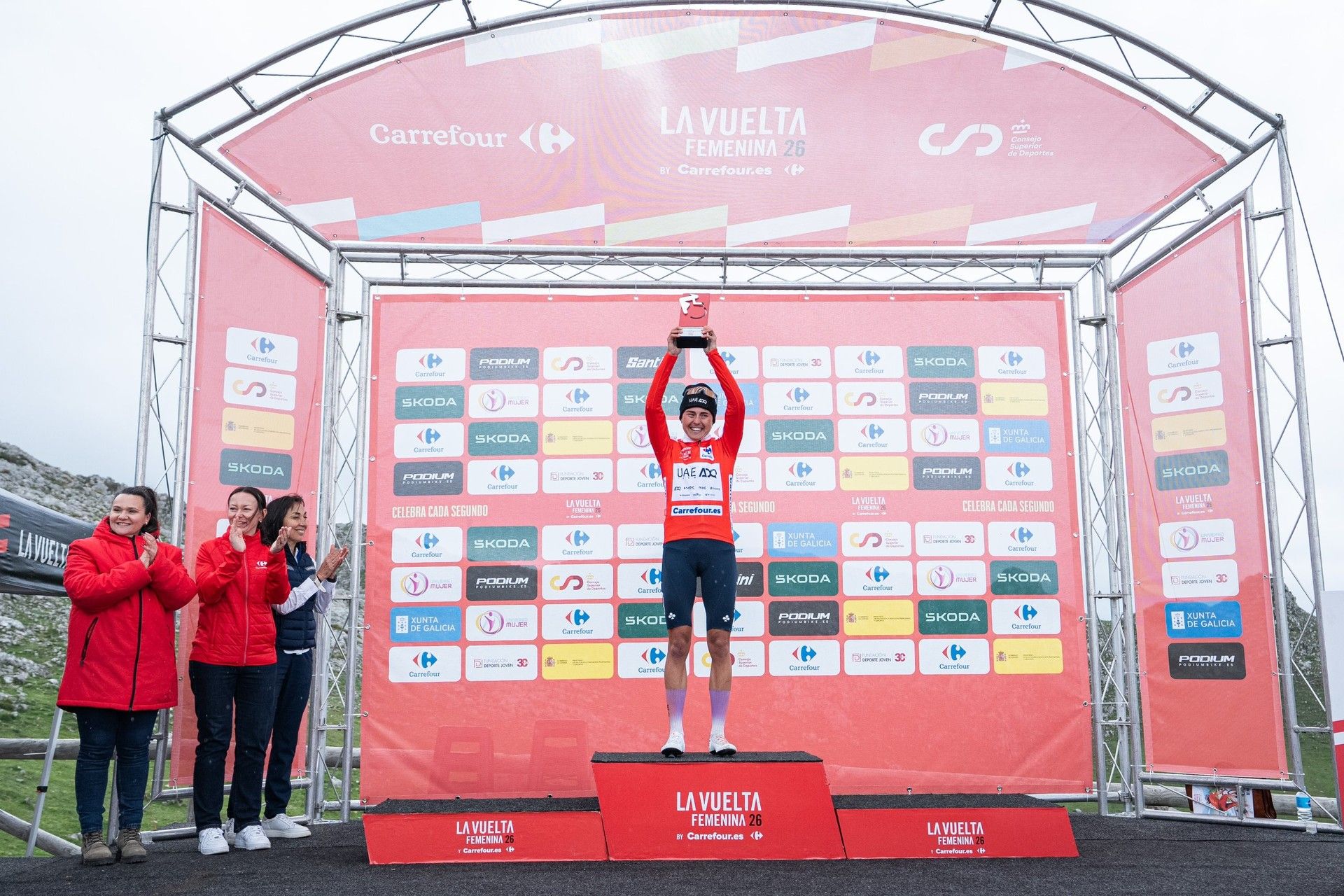 Paula Blasi stands on the La Vuelta Femenina podium after Stage 7 to L’Angliru, with two people in red jackets beside it