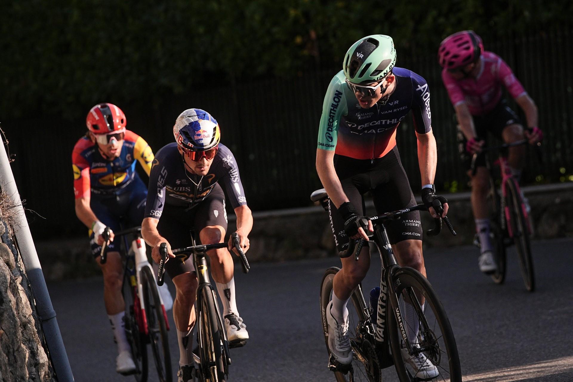 Three professional cyclists ride side by side on a road, wearing helmets and team racing kits.