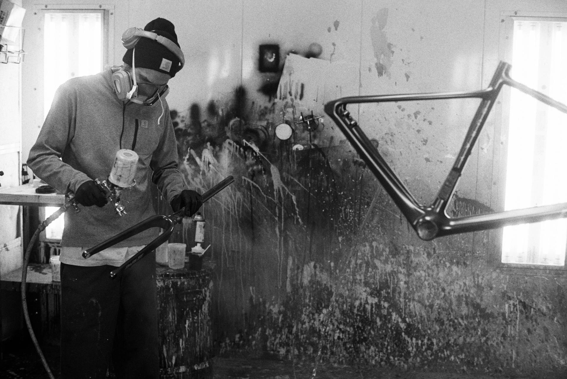 Black and white image of a craftsperson working on a bicycle frame at their bench, the frame held in a stand as they attend to joint details.
